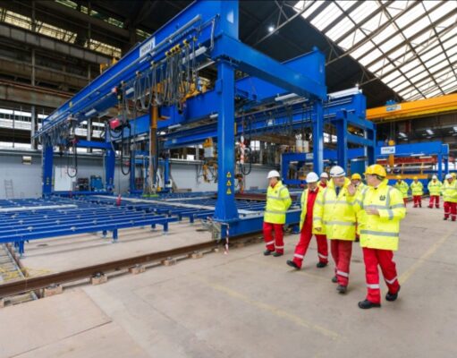 Group of workers in yellow high-visibility jackets and white hard hats walking beside a large blue overhead crane in an industrial factory setting with rails on the floor.