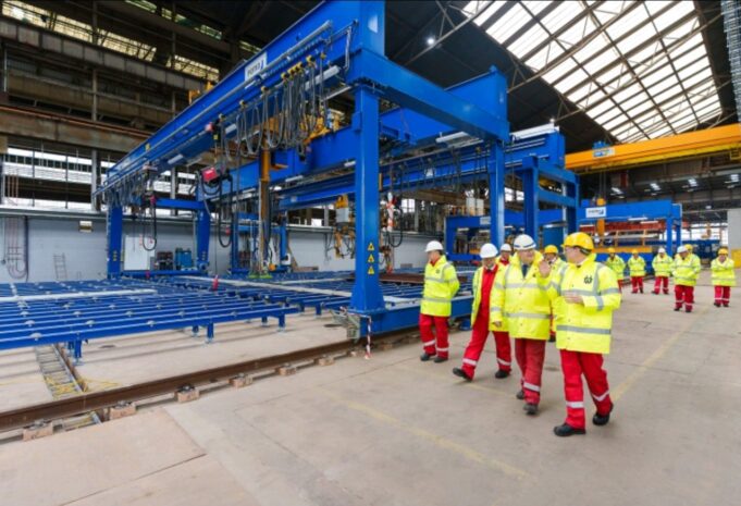 Group of workers in yellow high-visibility jackets and white hard hats walking beside a large blue overhead crane in an industrial factory setting with rails on the floor.