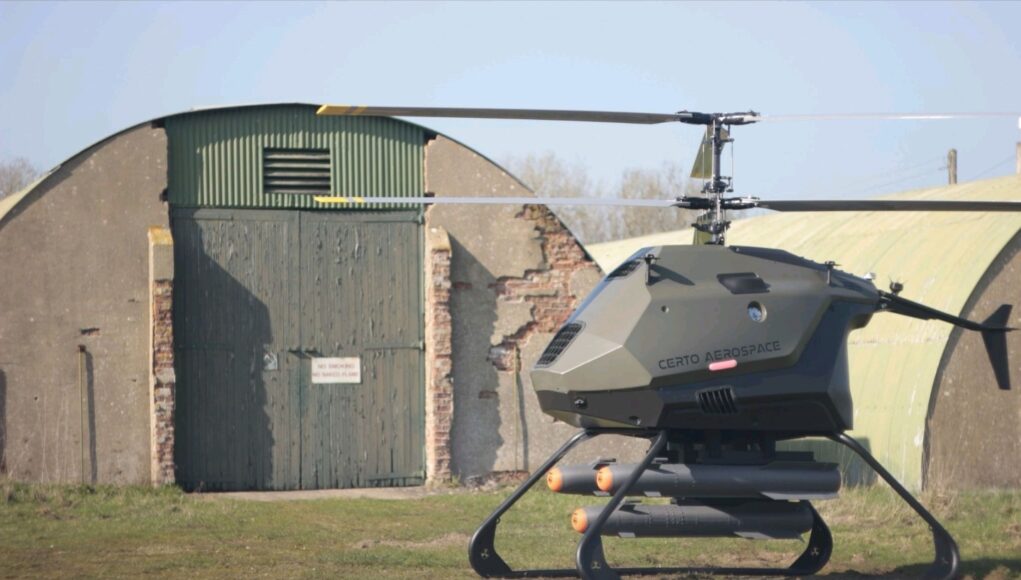 Futuristic gray helicopter drone with landing skids, labeled Certo Aerospace, parked in a grassy field beside a weathered hangar.