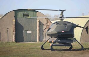 Futuristic gray helicopter drone with landing skids, labeled Certo Aerospace, parked in a grassy field beside a weathered hangar.