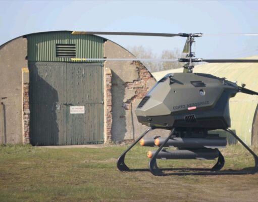 Futuristic gray helicopter drone with landing skids, labeled Certo Aerospace, parked in a grassy field beside a weathered hangar.