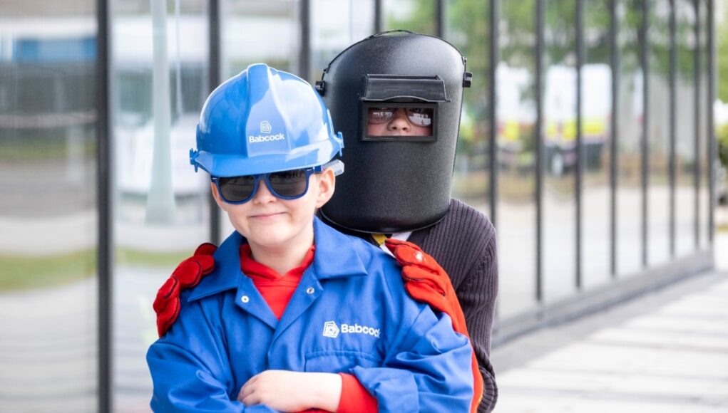 Young child in a blue hard hat and jacket with safety gloves stands in front of a person wearing a welding mask outdoors near a glass building.