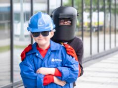 Young child in a blue hard hat and jacket with safety gloves stands in front of a person wearing a welding mask outdoors near a glass building.