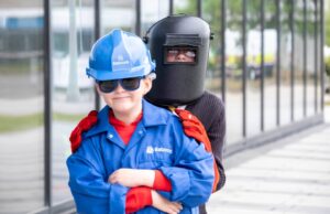Young child in a blue hard hat and jacket with safety gloves stands in front of a person wearing a welding mask outdoors near a glass building.