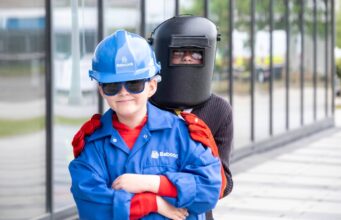 Young child in a blue hard hat and jacket with safety gloves stands in front of a person wearing a welding mask outdoors near a glass building.