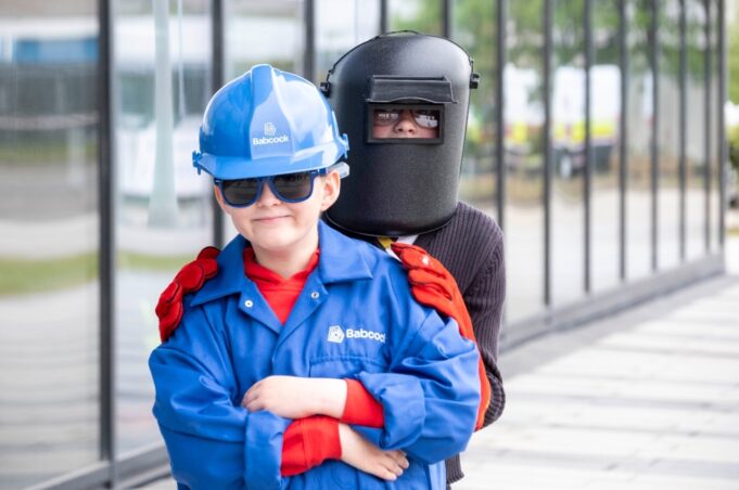 Young child in a blue hard hat and jacket with safety gloves stands in front of a person wearing a welding mask outdoors near a glass building.