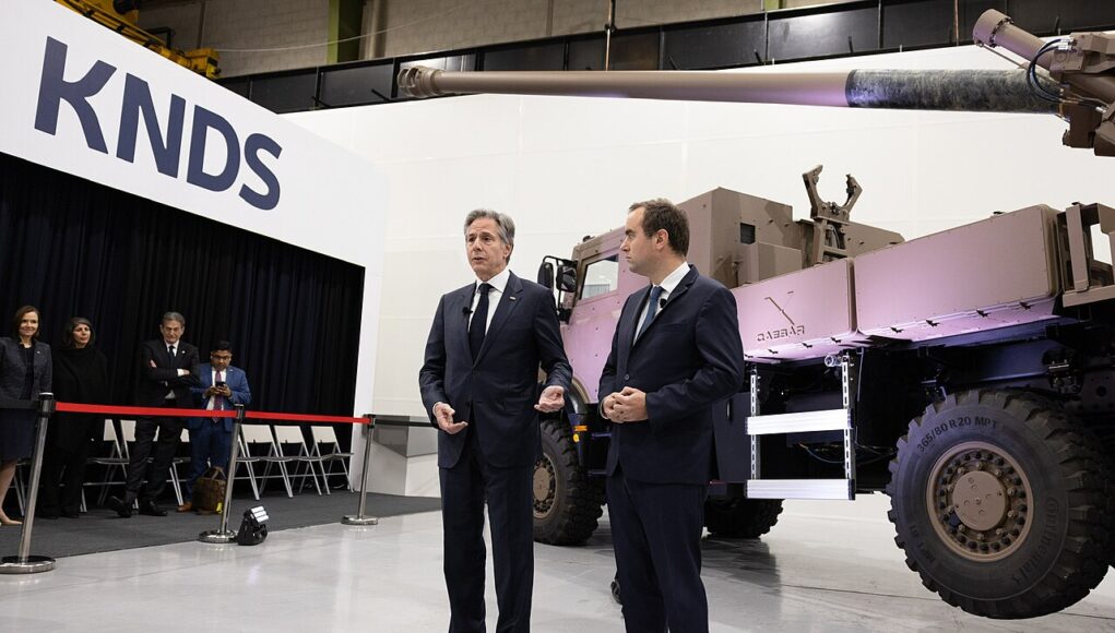 Two men in dark suits speak to an audience beside a large military vehicle on display at a KNDS exhibition, with a red rope barrier in front and people watching in the background.