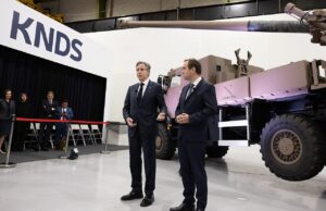 Two men in dark suits speak to an audience beside a large military vehicle on display at a KNDS exhibition, with a red rope barrier in front and people watching in the background.
