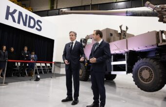 Two men in dark suits speak to an audience beside a large military vehicle on display at a KNDS exhibition, with a red rope barrier in front and people watching in the background.