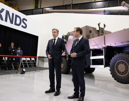 Two men in dark suits speak to an audience beside a large military vehicle on display at a KNDS exhibition, with a red rope barrier in front and people watching in the background.