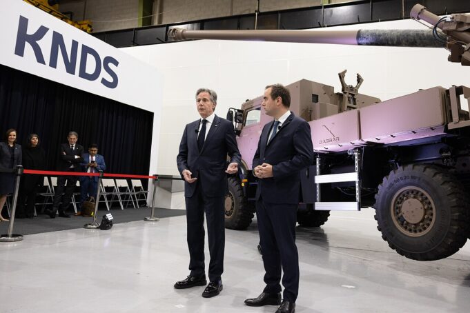 Two men in dark suits speak to an audience beside a large military vehicle on display at a KNDS exhibition, with a red rope barrier in front and people watching in the background.