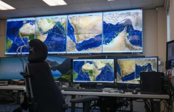 Mission control desk with multiple monitors displaying satellite maps of the Middle East and Indian Ocean; a high-backed chair in front of the setup.