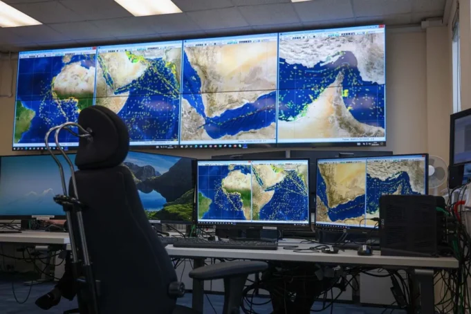 Mission control desk with multiple monitors displaying satellite maps of the Middle East and Indian Ocean; a high-backed chair in front of the setup.
