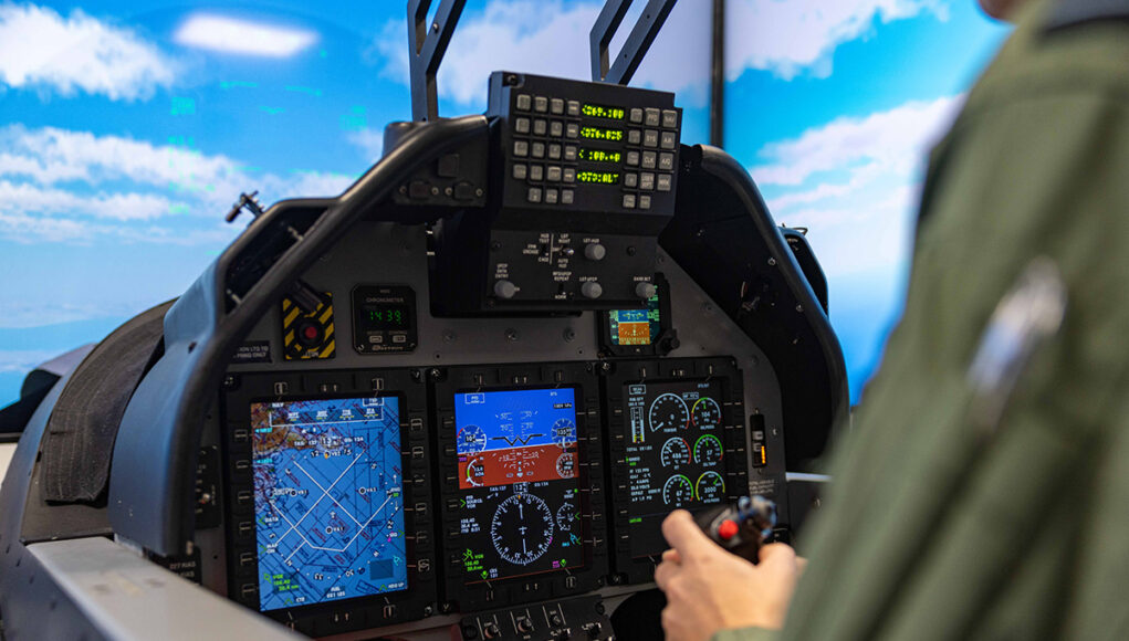 Pilot in a cockpit operating flight controls with touchscreen displays and avionics visible ahead of him to the blue sky outside the canopy.