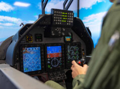 Pilot in a cockpit operating flight controls with touchscreen displays and avionics visible ahead of him to the blue sky outside the canopy.