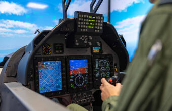 How simulation is speeding up the RAF pilot pipeline Pilot in a cockpit operating flight controls with touchscreen displays and avionics visible ahead of him to the blue sky outside the canopy.