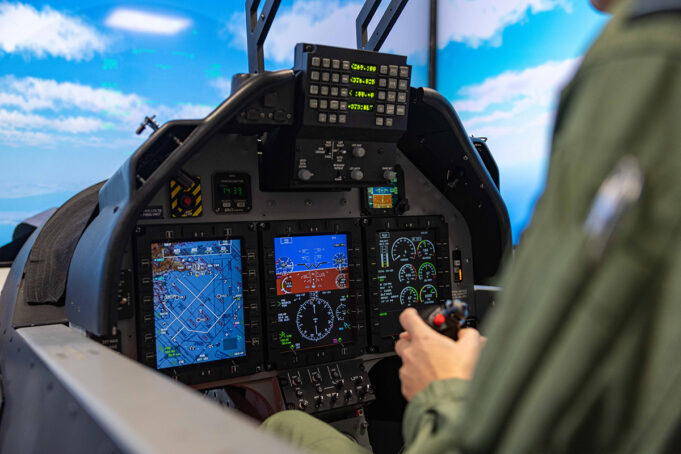 Pilot in a cockpit operating flight controls with touchscreen displays and avionics visible ahead of him to the blue sky outside the canopy.