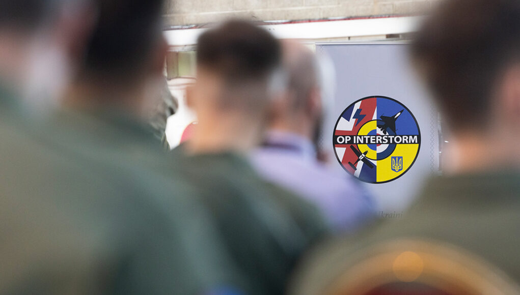 WIT-20260429-0115-IS_Super_Grad-066 Circular patch reading 'OP INTERSTORM' with blue, red, and yellow segments, seen among blurred soldiers in the foreground.
