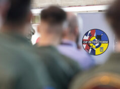 Circular patch reading 'OP INTERSTORM' with blue, red, and yellow segments, seen among blurred soldiers in the foreground.