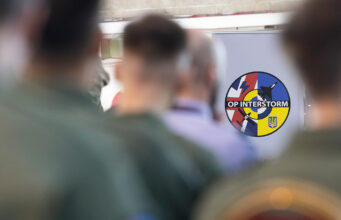 Circular patch reading 'OP INTERSTORM' with blue, red, and yellow segments, seen among blurred soldiers in the foreground.