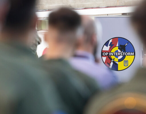 Ukrainian pilots graduate from British flying programme Circular patch reading 'OP INTERSTORM' with blue, red, and yellow segments, seen among blurred soldiers in the foreground.
