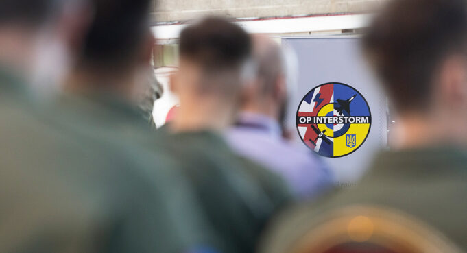 Circular patch reading 'OP INTERSTORM' with blue, red, and yellow segments, seen among blurred soldiers in the foreground.