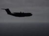 Silhouette of a four-engine military transport aircraft (C-130-like) flying over the ocean at dusk with a cloudy sky.