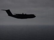 Silhouette of a four-engine military transport aircraft (C-130-like) flying over the ocean at dusk with a cloudy sky.