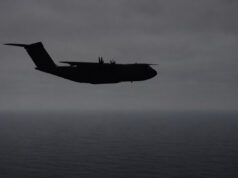 Silhouette of a four-engine military transport aircraft (C-130-like) flying over the ocean at dusk with a cloudy sky.