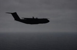 Silhouette of a four-engine military transport aircraft (C-130-like) flying over the ocean at dusk with a cloudy sky.