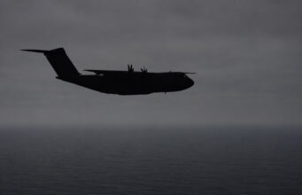 Silhouette of a four-engine military transport aircraft (C-130-like) flying over the ocean at dusk with a cloudy sky.