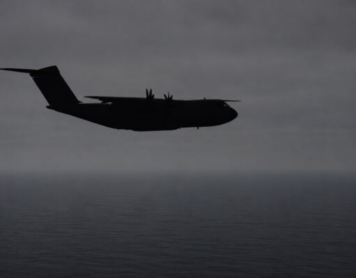 Silhouette of a four-engine military transport aircraft (C-130-like) flying over the ocean at dusk with a cloudy sky.