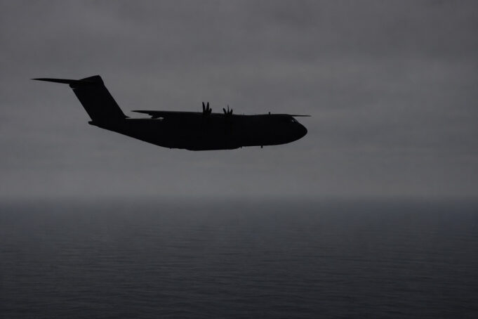 Silhouette of a four-engine military transport aircraft (C-130-like) flying over the ocean at dusk with a cloudy sky.