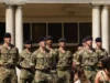 Group of soldiers in camouflage uniforms standing at attention in front of a columned building outdoors.