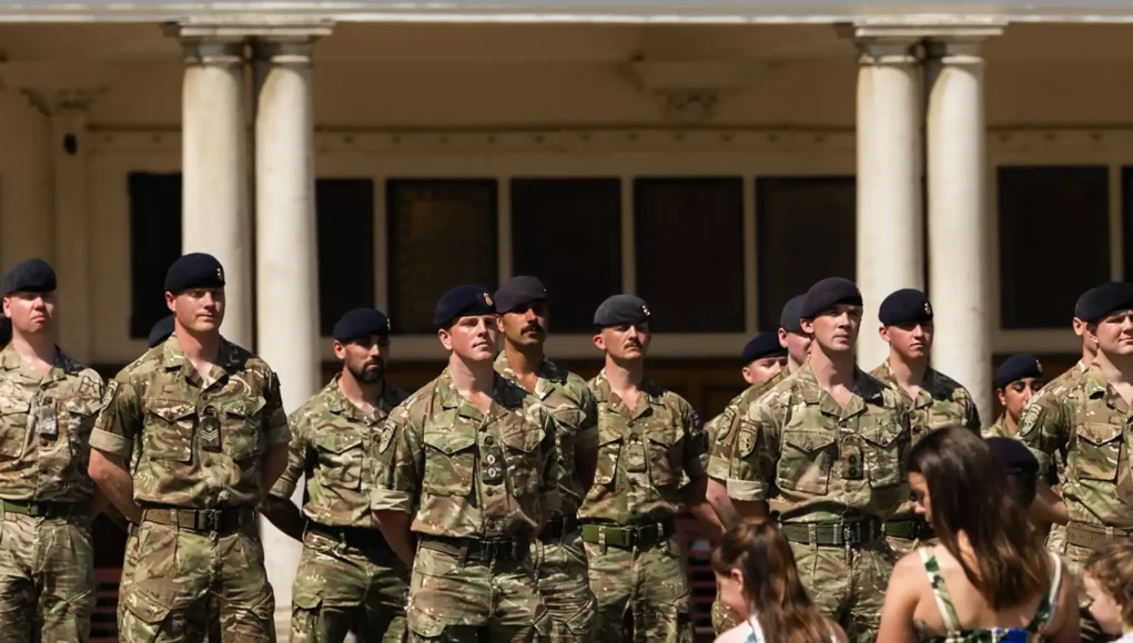 Group of soldiers in camouflage uniforms standing at attention in front of a columned building outdoors.