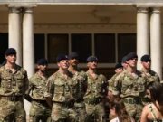 Group of soldiers in camouflage uniforms standing at attention in front of a columned building outdoors.