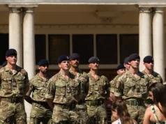 Group of soldiers in camouflage uniforms standing at attention in front of a columned building outdoors.