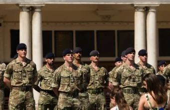 Group of soldiers in camouflage uniforms standing at attention in front of a columned building outdoors.