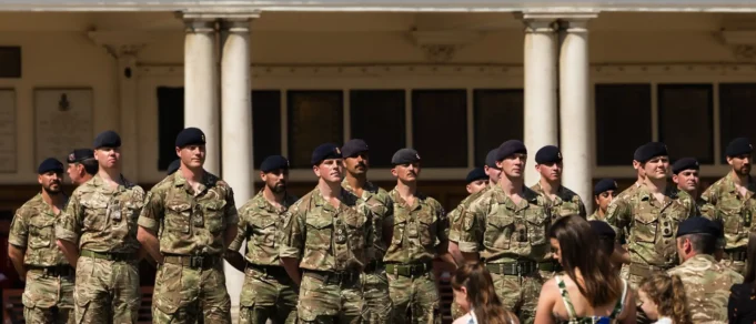 Group of soldiers in camouflage uniforms standing at attention in front of a columned building outdoors.