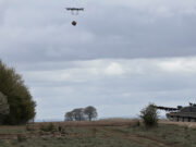 Quadcopter drone carrying a payload over a field with a military tank on the right under cloudy skies