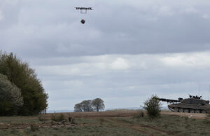 Quadcopter drone carrying a payload over a field with a military tank on the right under cloudy skies
