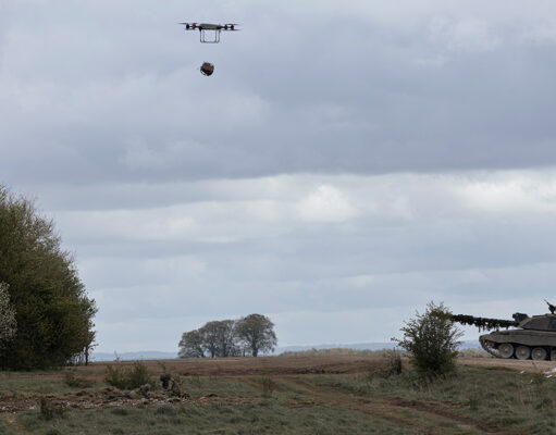 Quadcopter drone carrying a payload over a field with a military tank on the right under cloudy skies