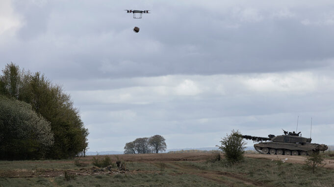 Quadcopter drone carrying a payload over a field with a military tank on the right under cloudy skies