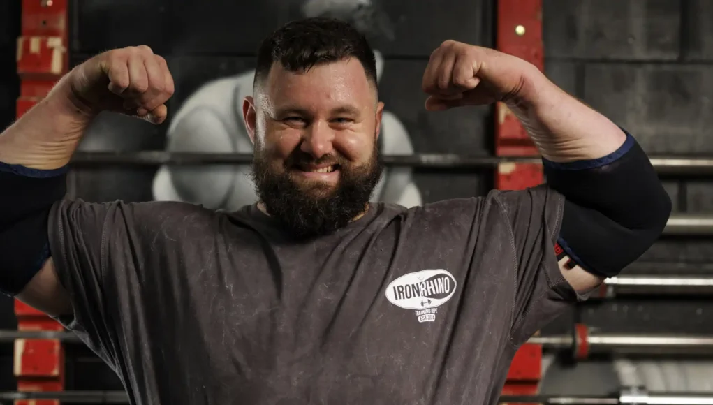 ahqsgt1-official-20260551 Bearded man in a gym flexing his biceps, wearing an Iron Rhino t-shirt, with weight racks in the background.
