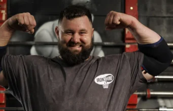 Bearded man in a gym flexing his biceps, wearing an Iron Rhino t-shirt, with weight racks in the background.