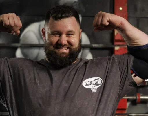 Bearded man in a gym flexing his biceps, wearing an Iron Rhino t-shirt, with weight racks in the background.