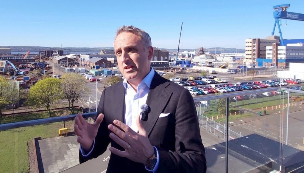 Man in a dark suit and lapel microphone speaks on a balcony with an industrial harbor and parking lot in the background.