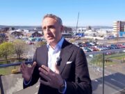 Man in a dark suit and lapel microphone speaks on a balcony with an industrial harbor and parking lot in the background.