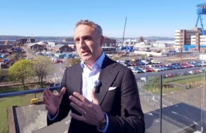 Man in a dark suit and lapel microphone speaks on a balcony with an industrial harbor and parking lot in the background.