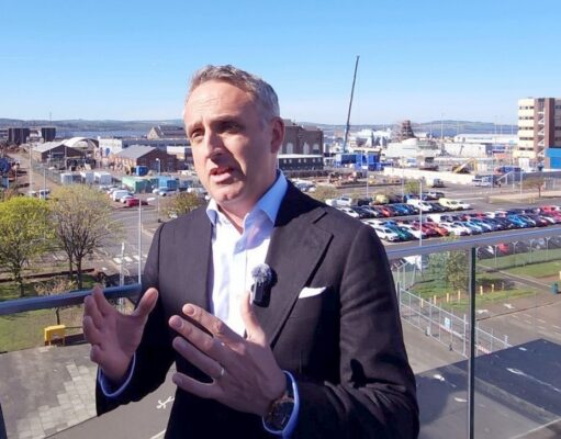 Man in a dark suit and lapel microphone speaks on a balcony with an industrial harbor and parking lot in the background.